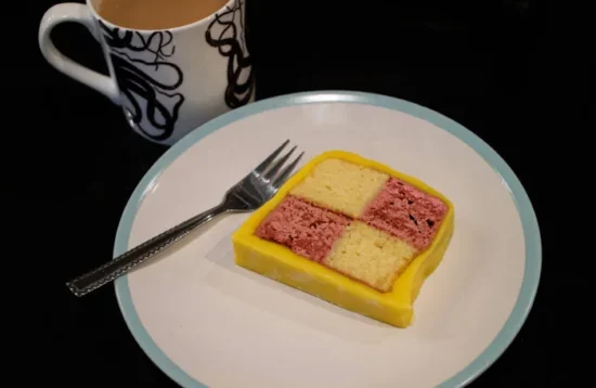 A slice of Battenberg cake on a saucer with a cup of tea in the background.