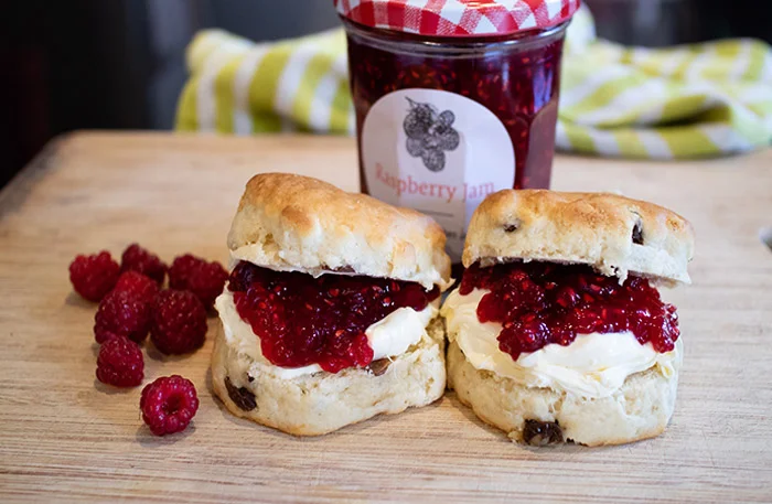 Two scones filled with raspberry jam and clotted cream. There is a jar of raspberry jam in the background and some fresh raspberries on the left side. 