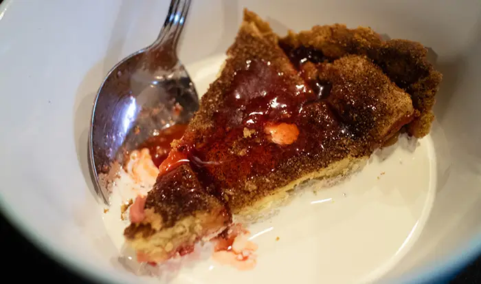A slice of the strawberry & gooseberry pie, in a bowl, with a spoon by the side. There is thick cream which has been poured into the bowl. 