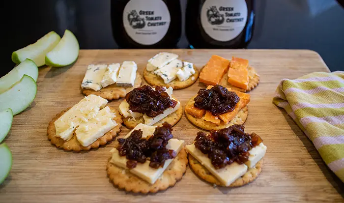 The Green Tomato Chutney being used on some cheese with crackers. All are on a wooden board with two jars of the chutney in the background. 